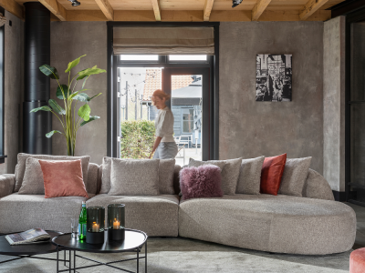 Moderne woonkamer met grote, grijze bank, kleurrijke kussens, ronde tafels en een vrouw die buiten langs het raam loopt.