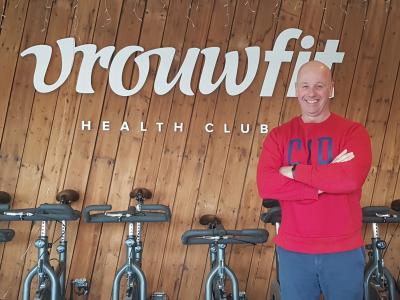Man in rode trui staat met gevouwen armen voor een wand met Vrouwfit Health Club logo en fitnessfietsen.