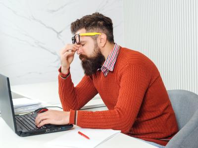 Man in rode trui met bril wrijft vermoeid zijn ogen terwijl hij aan een laptop werkt.