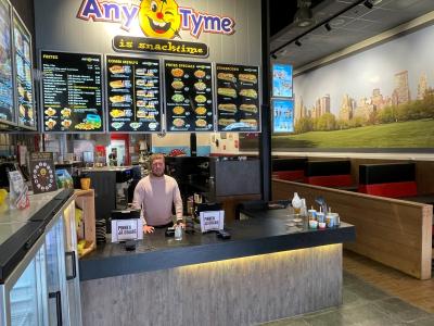Arjan Holwerda staat achter de balie in een AnyTyme snackbar met menuborden en zitgedeelte op de achtergrond.
