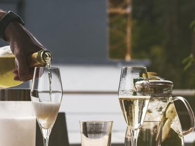Een hand schenkt mousserende wijn in een glas bij een tafel met glazen en een schenkkan met ijs en schijfjes fruit.