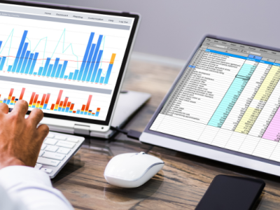 Twee computerschermen tonen grafieken en spreadsheets terwijl een persoon gegevens analyseert met een toetsenbord en muis.