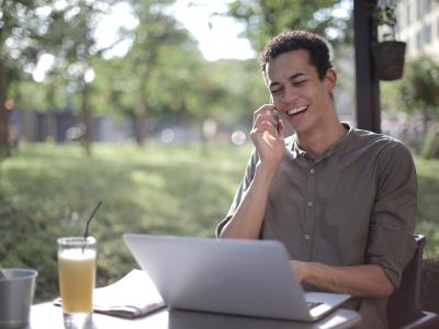 Een man werkt lachend op zijn laptop in een zonnige tuin, terwijl hij telefoon oortje gebruikt.