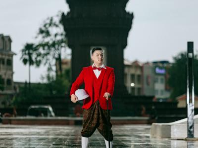 Een man in een rood kostuum houdt een witte helm vast en staat voor een architectonisch monument.