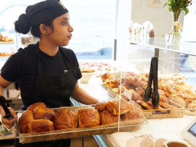Medewerker bij Le Pain Quotidien met een dienblad vol versgebakken croissants en gebak in een bakkerijomgeving.
