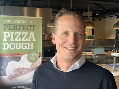 Man glimlacht voor een poster over pizzadeeg in een restaurantkeuken met laptops en kookgerei op de achtergrond.