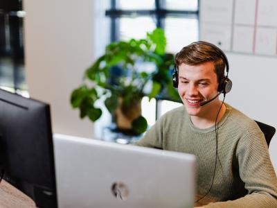 Man met headset werkt glimlachend aan een bureau met computer, plant en whiteboard op de achtergrond.