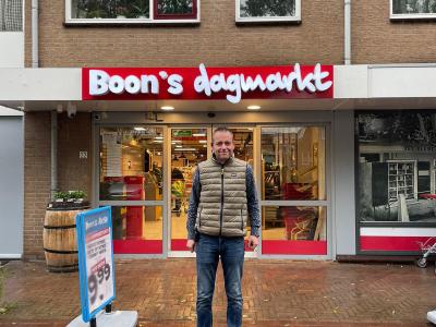 Man in casual kleding staat voor de ingang van Boon's dagmarkt met een reclamebord op de voorgrond.