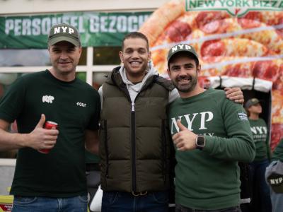 Drie mannen in NYP-kleding poseren lachend voor een New York Pizza achtergrond met daarop een personeelsbanner.