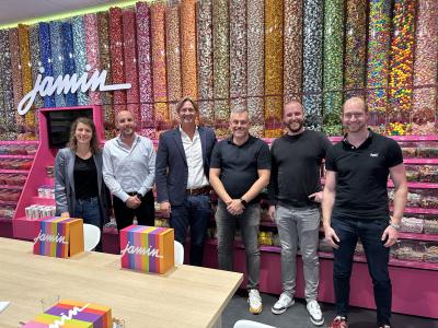 Zes mensen poseren voor kleurrijke snoepwanden in een Jamin-winkel, naast tafels met logo-dozen.
