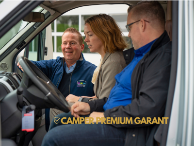 Drie mensen zitten in een camper terwijl een man een koppel het stuur en interieur laat zien.