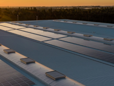 Zonnepanelen op het dak van een industrieel gebouw bij zonsondergang met bomen aan de horizon.