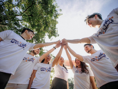 Teamleden in witte shirts staan in een kring en maken een gezamenlijke high five onder een heldere hemel.