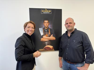 Twee mensen staan voor een poster van Gelderblom Slagers met een afbeelding van een slager.