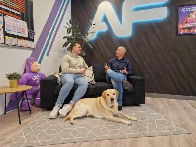 Twee mannen in gesprek op een bank in een kantooromgeving met een hond op de vloer en een groot logo op de muur.