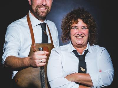 Twee personen in slagerskleding bij een houten snijplank met een groot stuk rauw vlees en een mes.