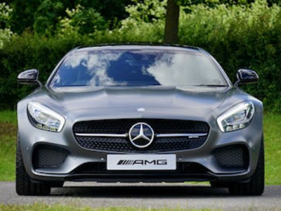 Grijze luxe sportauto met AMG-logo geparkeerd op een weg met groene struiken op de achtergrond.