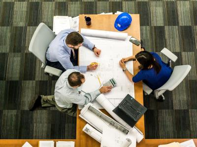Drie mensen in overleg rond een tafel met bouwtekeningen en een laptop, doelen bespreken op kantooromgeving.
