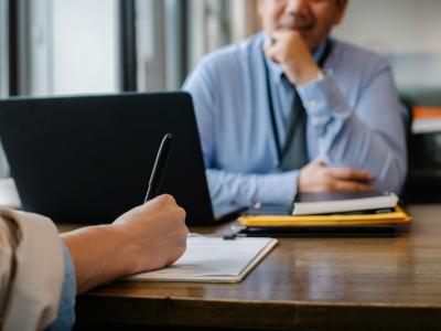 Twee zakenmensen aan een tafel, waarvan één schrijft in een document met een pen naast een open laptop.