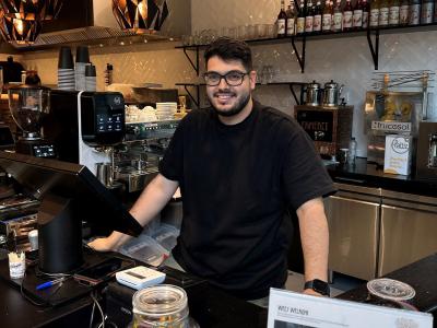 Jonge ondernemer achter de toonbank van zijn restaurant met koffieapparatuur en voorraad op de achtergrond.