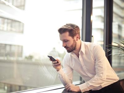 Man in witte blouse kijkt geconcentreerd op smartphone bij groot raam in modern kantoor.