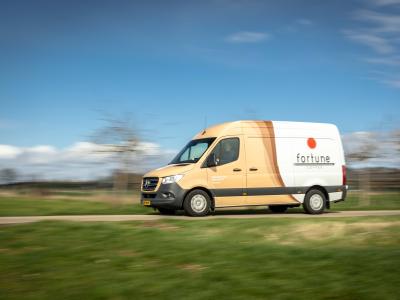 Een rijdende bestelwagen van Fortune Coffee op een weg omgeven door gras en bomen.