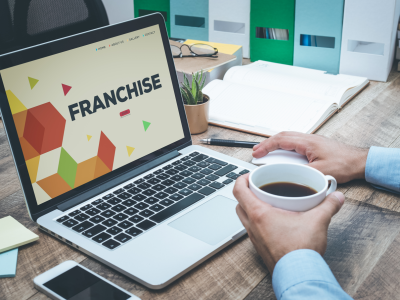 Laptop met franchisewebsite op scherm, omringd door kantoorbenodigdheden en een persoon met een kop koffie.