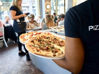 Medewerker serveert pizza's in een druk bezocht buurtrestaurant met etende gasten op de achtergrond.
