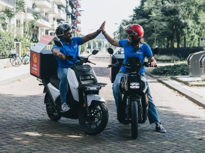 Twee bezorgers op scooters geven elkaar een high-five op een zonnige straat.
