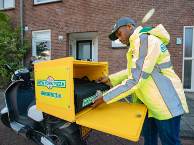 Een bezorger van New York Pizza pakt een thermobox uit de bezorgscooter voor de deur van een bakstenen huis.