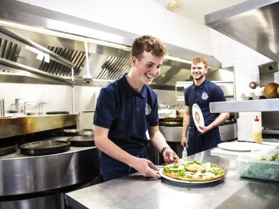 Twee lachende medewerkers in de keuken bereiden een pannenkoek voor bij De Pannekoekenbakker.