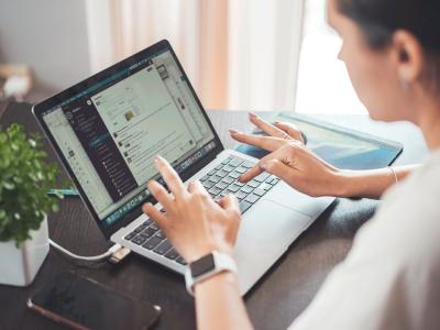 Persoon werkt op een laptop waarin een chatapplicatie openstaat, met op de achtergrond een smartphone en een plant.