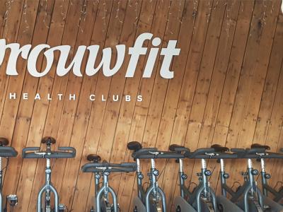 Een man in rode trui poseert voor het logo van Vrouwfit Health Clubs, omringd door hometrainers op een houten achtergrond.