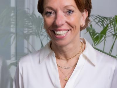 Een vrouw glimlacht naar de camera, gekleed in een witte blouse, met een groene plant op de achtergrond.