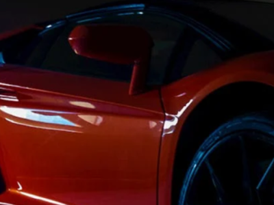 Close-up van een sportauto met oranje carrosserie en glanzende zwarte velgen, geparkeerd in een garageomgeving.
