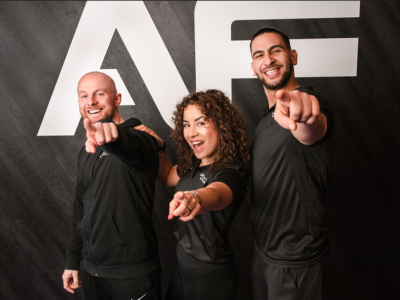 Drie lachende mensen in zwarte sportkleding wijzen naar de camera voor een grote AF-achtergrond.
