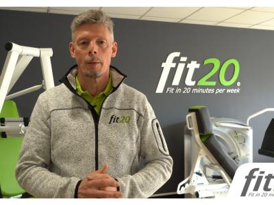 Man in fit20-kleding presenteert voor fitnessapparatuur met het fit20-logo op de achtergrond.