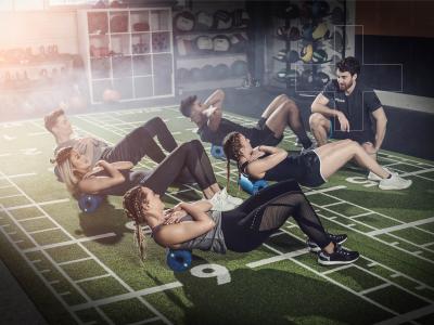 Groep mensen doet buikspieroefeningen op schuimrollers onder begeleiding van een trainer in een fitnessruimte.
