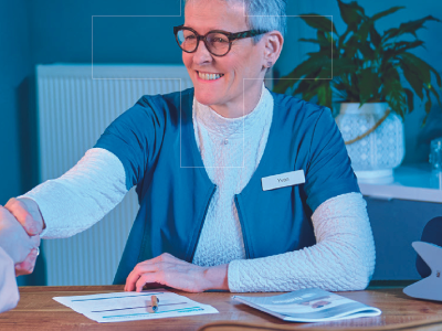 Vrouw in blauwe jas schudt hand boven bureau met documenten en planten op de achtergrond.