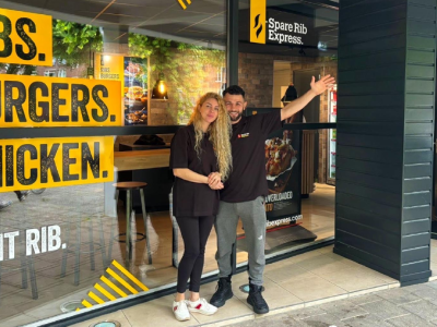 Twee mensen staan glimlachend voor de ingang van Spare Rib Express Weert, omringd door ramen met kleurrijke promotieteksten.