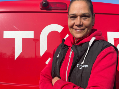 Persoon in rode kleding met zwart vest, glimlachend voor een rood voertuig met het logo van TOPP.