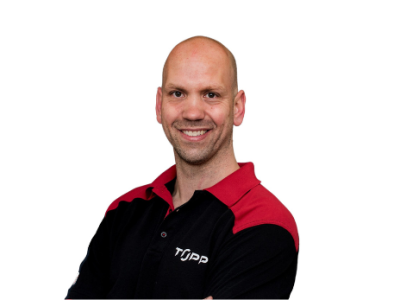 Man in zwart-rood TOPP-uniform staat glimlachend met gekruiste armen voor een witte achtergrond.