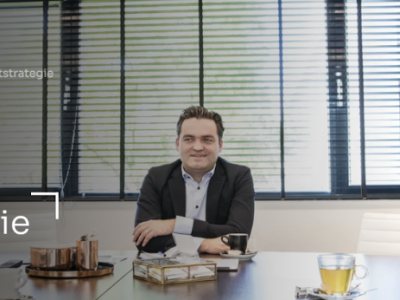 Vergadering in een kantoor met drie personen aan tafel, glazen water en thee, en zonlicht dat door jaloezieën schijnt.