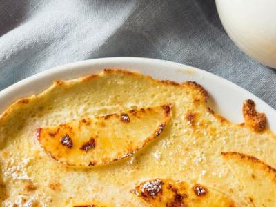 Twee pannenkoeken met poedersuiker en appels op een tafel, naast een rode appel en een melkkannetje.