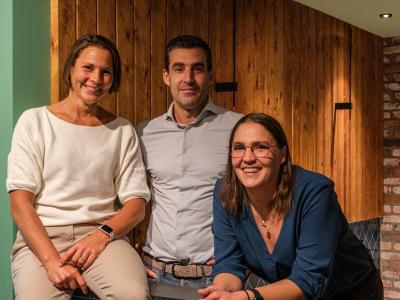 Drie lachende mensen poseren voor een houten en bakstenen muur in een warme, gezellige ruimte.