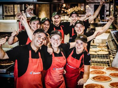 Een groep glimlachende Domino's-medewerkers poseert in de pizzeria, omringd door pizza's en ovens.