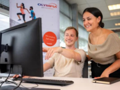 Twee mensen werken lachend samen achter een computer in een modern kantoor met een Olympia-poster op de achtergrond.