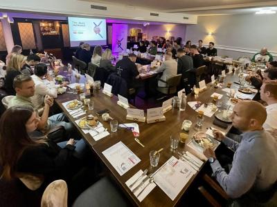 Een groep mensen zit aan een grote U-vormige tafel tijdens een zakelijke bijeenkomst met presentatiescherm.
