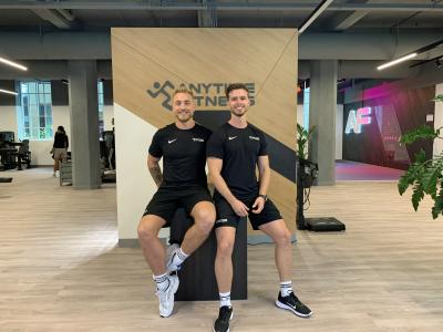 Twee mannen in sportkleding zitten voor een logo van Anytime Fitness in een moderne fitnessruimte.
