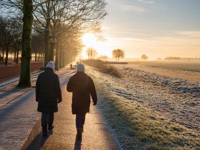 Twee personen in winterkleding lopen langs een besneeuwd pad bij zonsopgang in een landelijke omgeving.